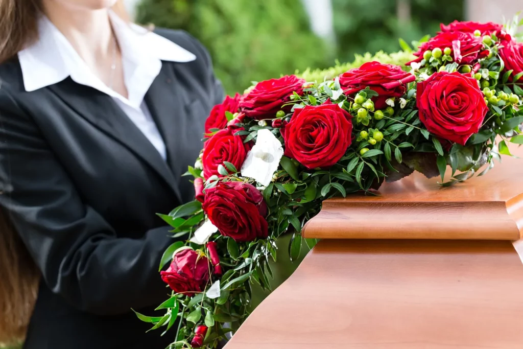 Femme en costume noir disposant une composition florale de roses rouges sur un cercueil en bois poli